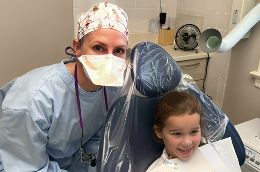 Pediatric dentist in surgical attire poses with young child patient in dental treatment room.