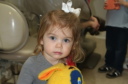 Young child with blonde hair and white bow holding colorful toy in dental office setting.