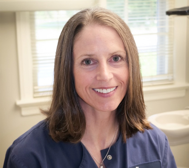 Professional headshot of smiling dental staff member in blue scrubs in clinical office setting.