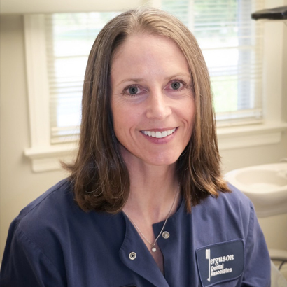 Professional female dentist smiling at camera in clinical dental office setting, wearing navy scrubs.