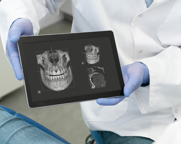 Dentist in gloves holding tablet displaying dental X-ray images of skull and teeth.
