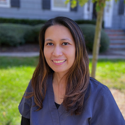 Professional headshot of a smiling woman in dark blue scrubs outdoors with residential background.