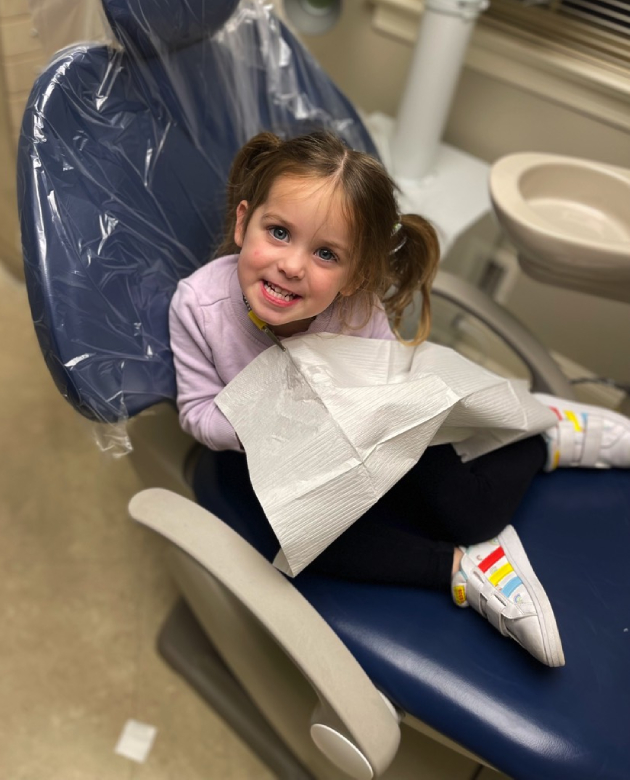 Young child in dental chair holding paper bib, smiling during dental visit.
