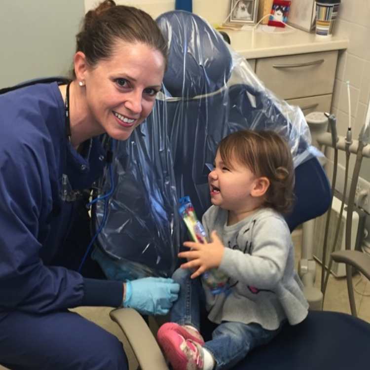 Dental professional in blue scrubs and gloves smiling with young toddler in dental office setting.