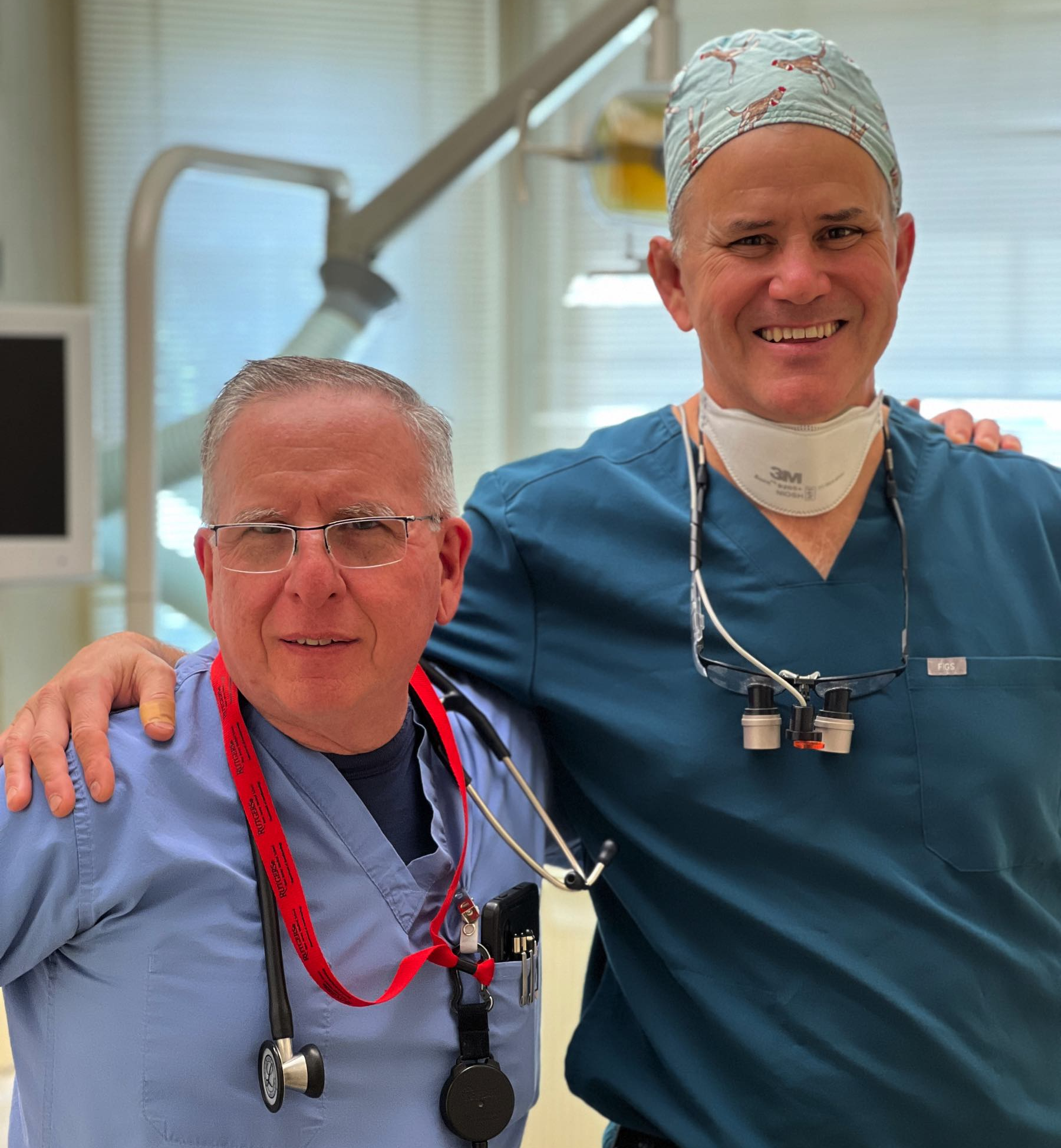 Two dental professionals posing together in clinical setting with surgical lights visible.