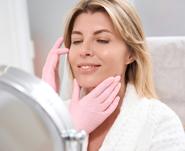 Woman applying pink towel to face in bathroom mirror during skincare routine.