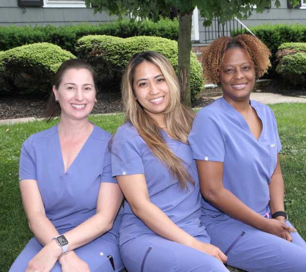 Three dental professionals in blue scrubs posing together outdoors in front of hedges.