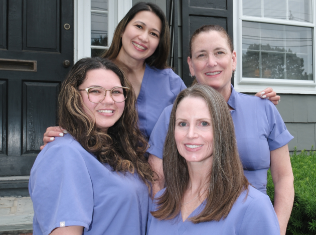 Four smiling dental professionals in blue scrubs pose together outside a brick dental office building.
