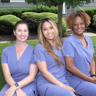 Three dental professionals in blue scrubs sitting outdoors in front of landscaping.