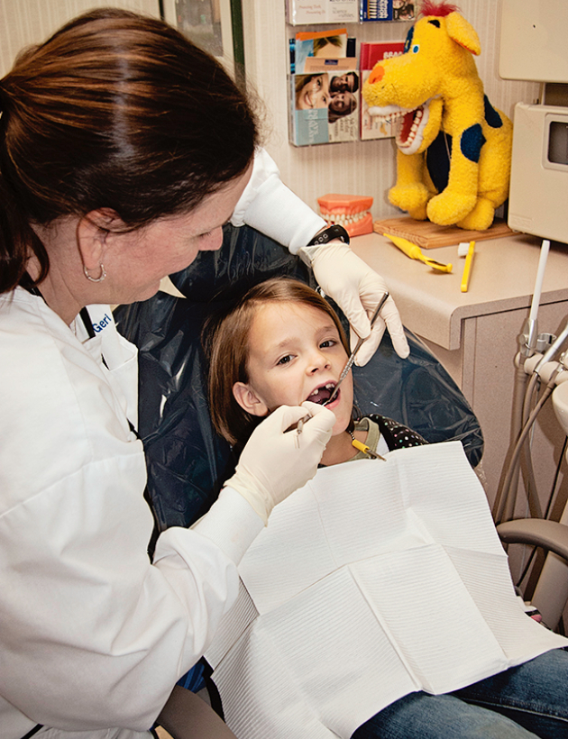 Pediatric dentist examining young child's teeth in dental office with yellow mascot decoration visible.