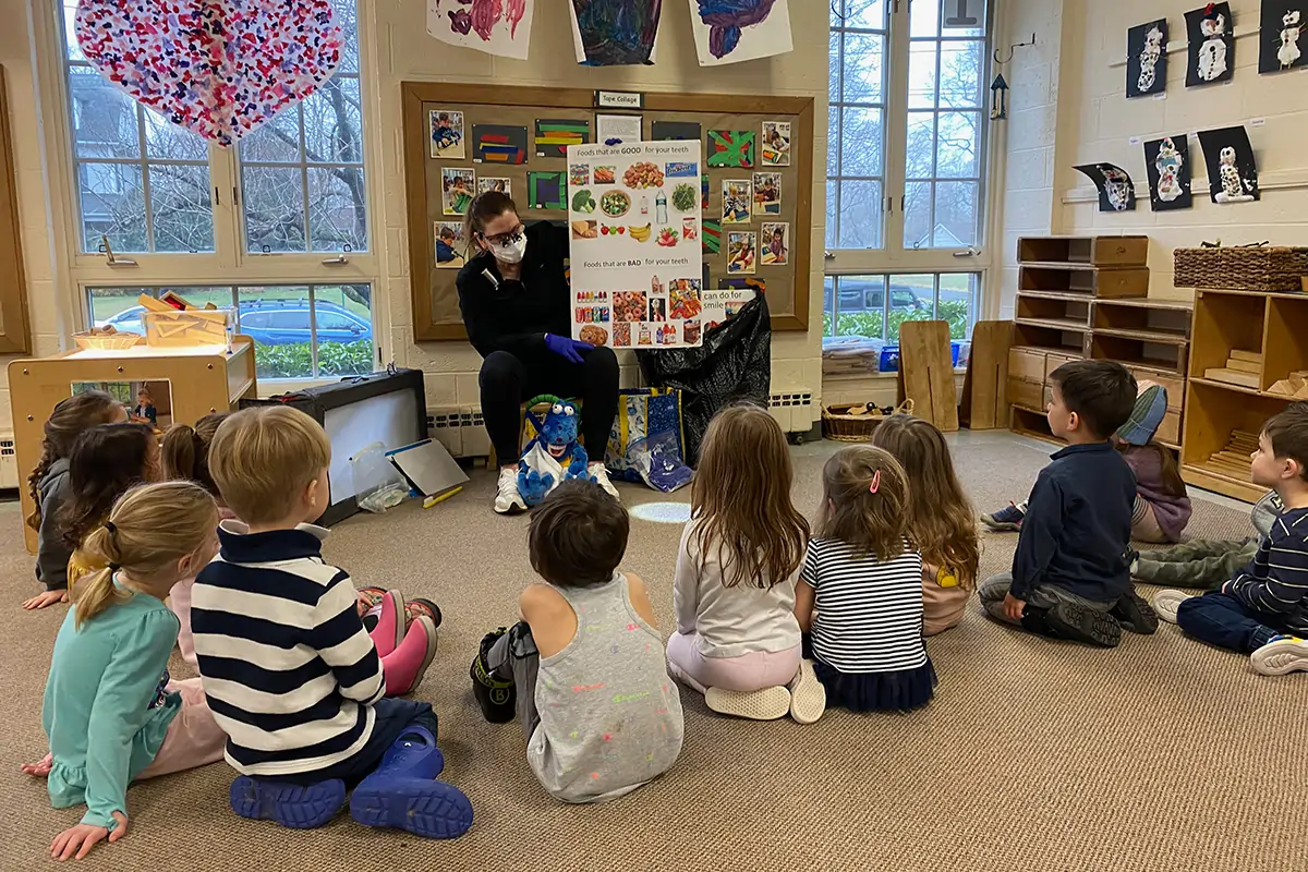Teacher reads to seated children in bright classroom with windows and educational displays.