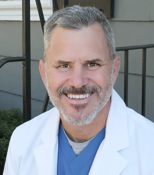 Male dentist in white coat and blue scrubs smiling at camera, professional headshot.