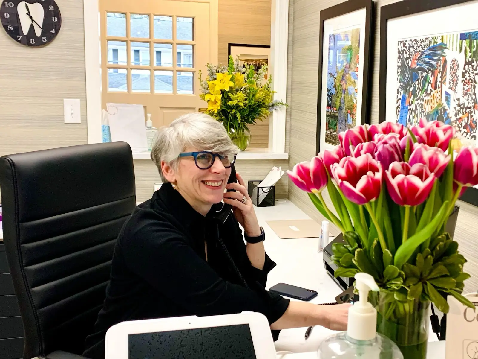 Dental professional seated at desk in modern office with pink tulips and computer visible.