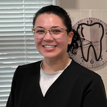 Smiling dental professional wearing glasses and black cardigan in office with clock on wall.