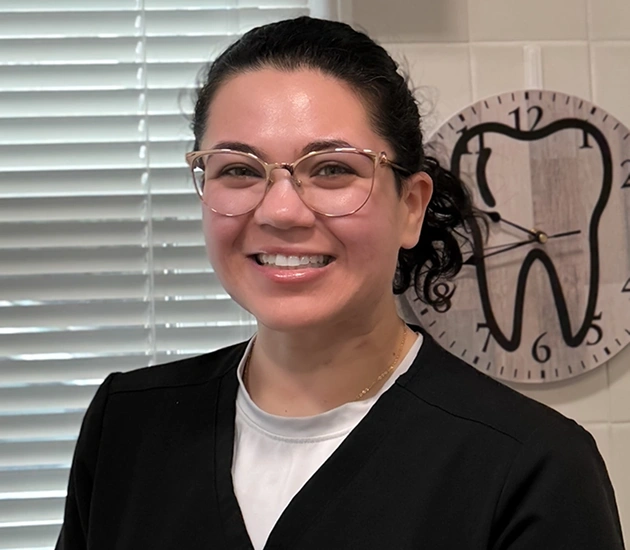 Smiling dental professional wearing glasses in office with tooth-shaped clock on wall.