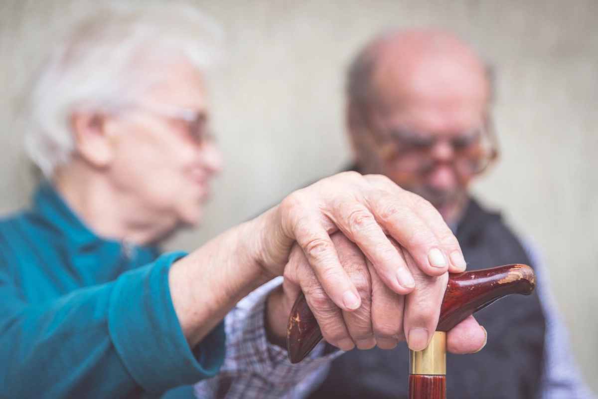 A close-up of an elderly woman's hand gently resting on top of an elderly man's hand as he grips a wooden walking cane, with both individuals softly blurred in the background.